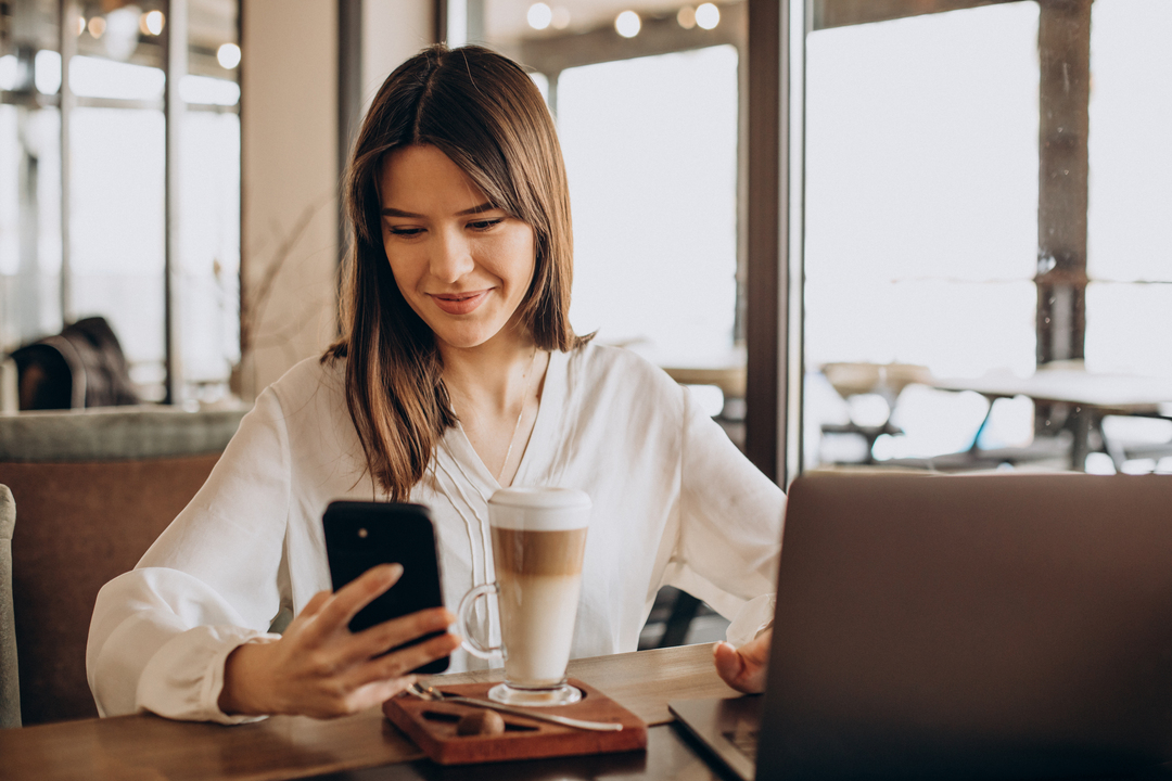 Imagem mostra mulher em café, mexendo no celular, enquanto faz networking.